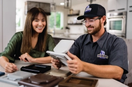 A homeowner and professional dryer vent cleaner sit in kitchen reviewing exterior dryer vent caps for replacement.